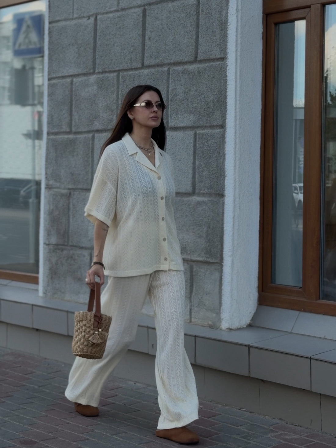 A woman stands outdoors on a sidewalk in front of a gray stone building, wearing a matching cream-colored two-piece lounge set consisting of a short-sleeve button-front shirt and wide-leg pants. She is also wearing brown shoes and carrying a small tan handbag.