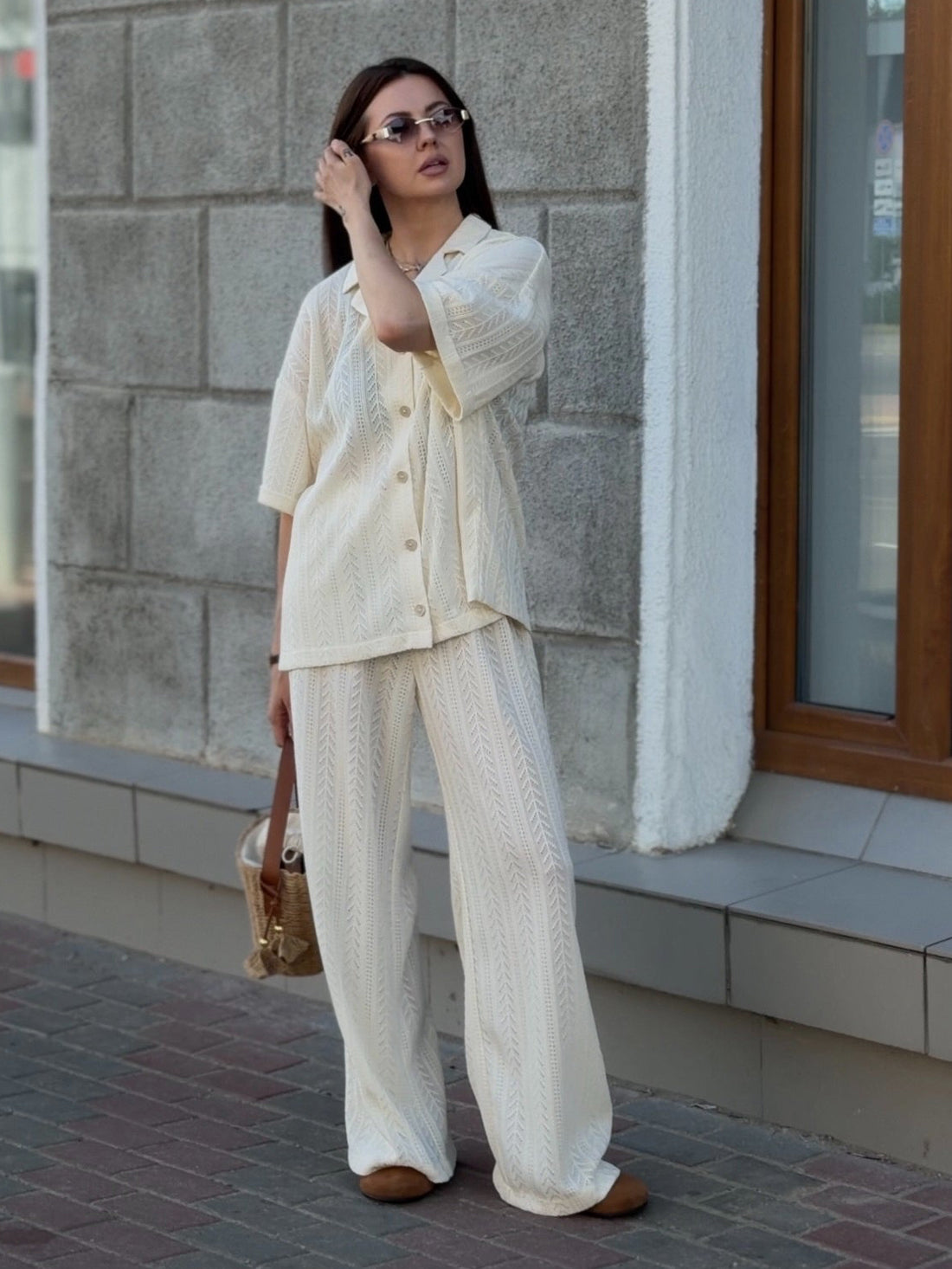A woman stands outdoors on a sidewalk in front of a gray stone building, wearing a matching cream-colored two-piece lounge set consisting of a short-sleeve button-front shirt and wide-leg pants. She is also wearing brown shoes and carrying a small tan handbag.